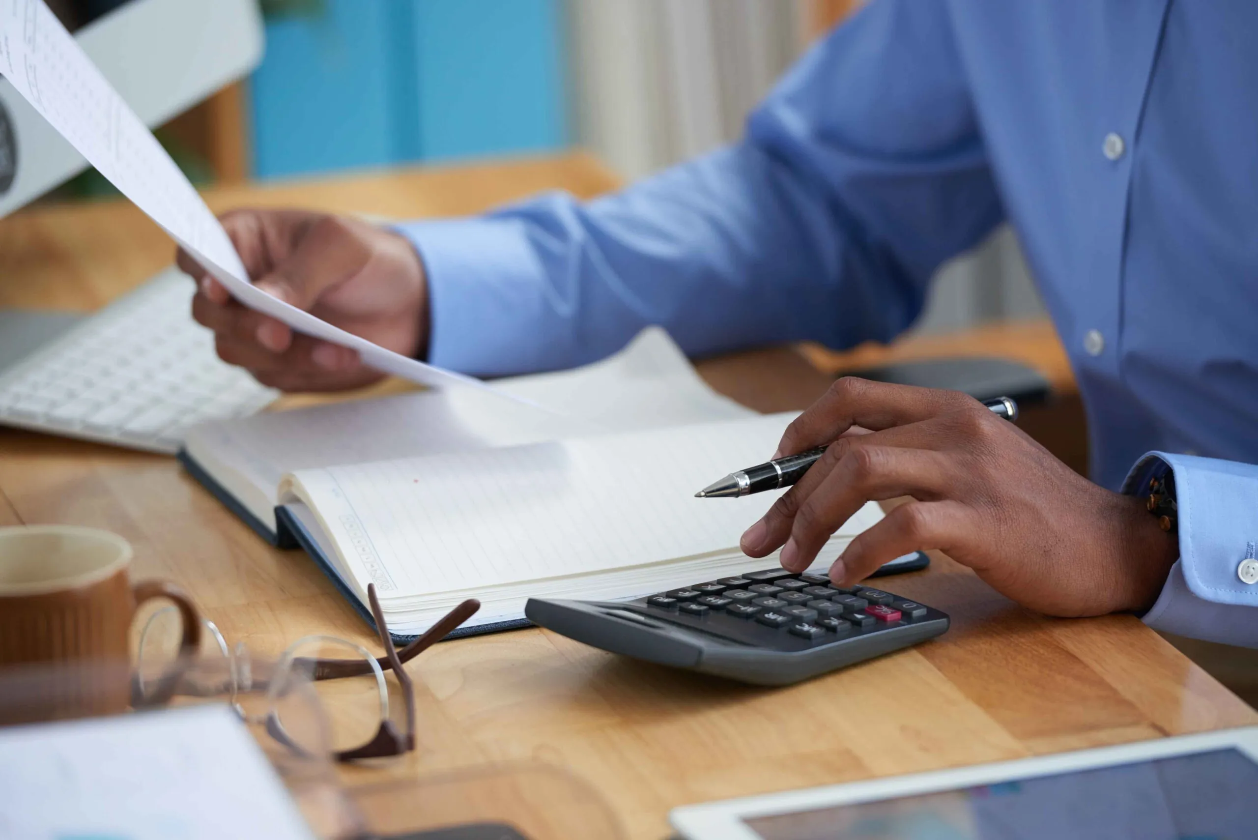 Person in a blue shirt holding a document and using a calculator beside an open notebook