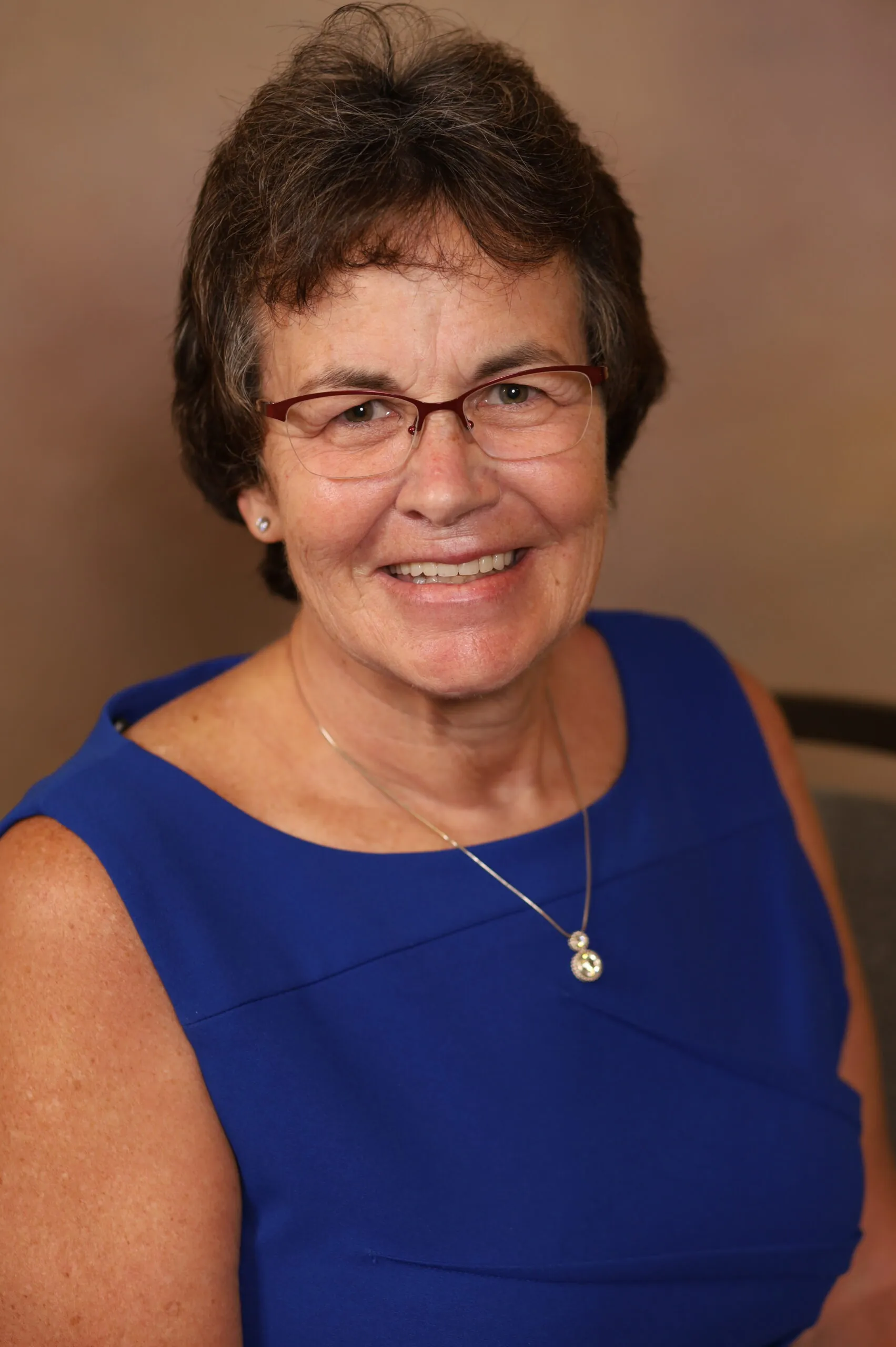 Smiling woman with short brown hair and red glasses wearing a blue sleeveless dress and pendant necklace