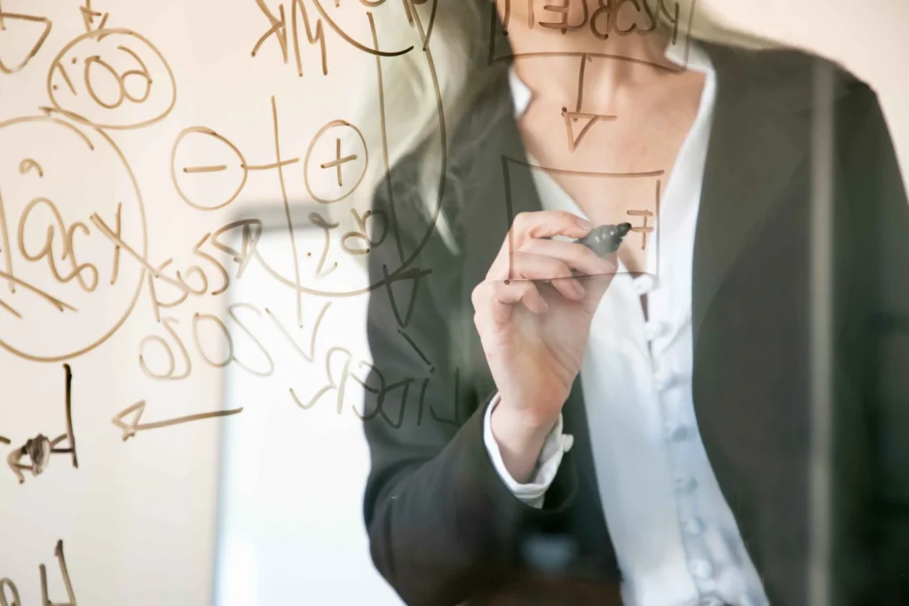Woman in a dark blazer writing formulas and diagrams on a glass board with a marker