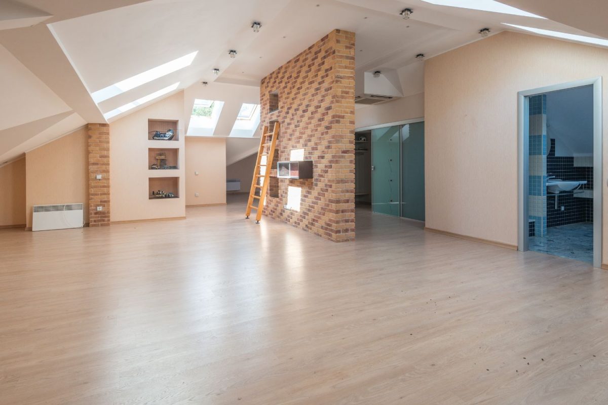 Empty attic apartment with hardwood floors, a brick accent wall with a ladder, skylights, and a view into a bathroom.