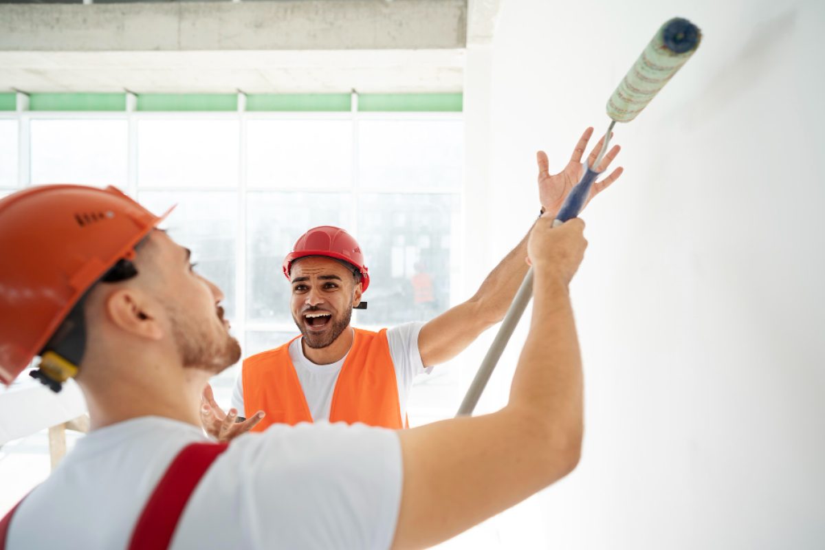 Two construction workers in hard hats discuss estimating paint coverage on a wall.